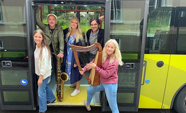 Fem smilende personer i døråpningen på en buss, en dame holder en harpe og en mann med saxofon