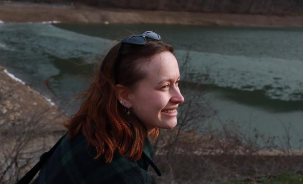 Kvinne med rødlig skulderlangt hår sitter fremfor det som ligner på en innsjø. Hun smiler og har solbriller på hodet. 