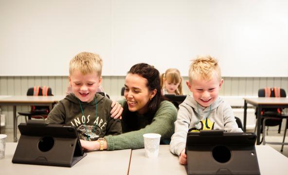 Lærer sitter på huk mellom to barneskoleelever i klasserom som har hver sin laptop på pulten