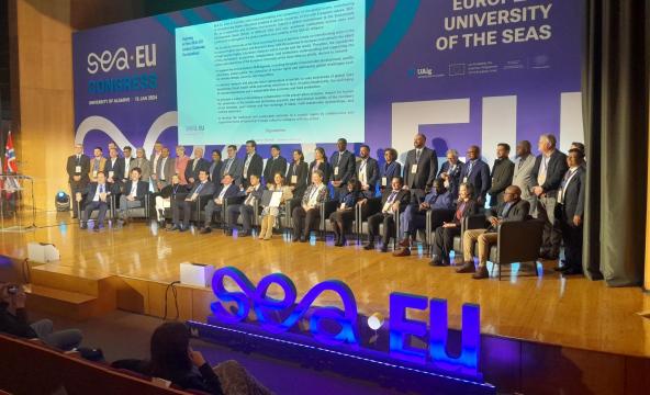 Mange mennesker sitter på en scene på en kongress. I forgrunnen et lysende SEA-EU-skilt