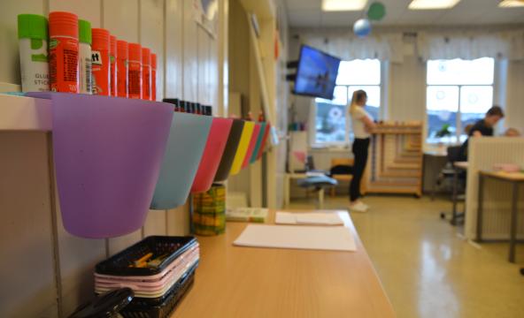 Klasserom barneskole med fargede kopper på vegg foran og lærer og elever i bakgrunnen. Foto