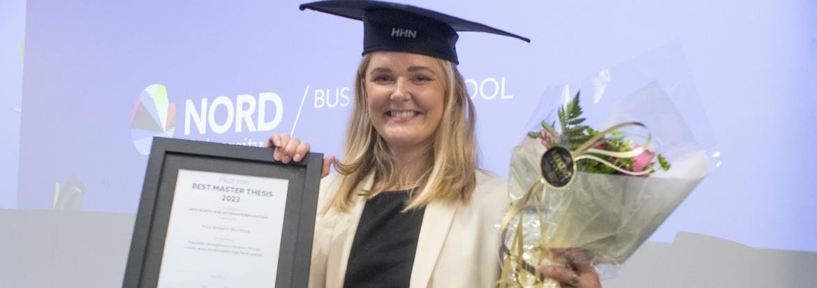 Female student, wearing a graduation hat, holding a diploma and flowers while smiling into the camera.