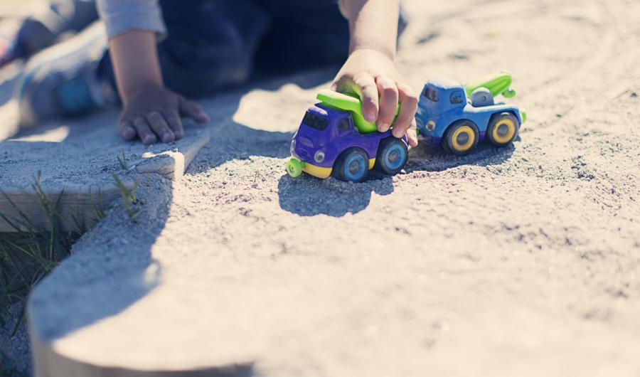 Et barn leker med en lekebil i ei sandkasse.