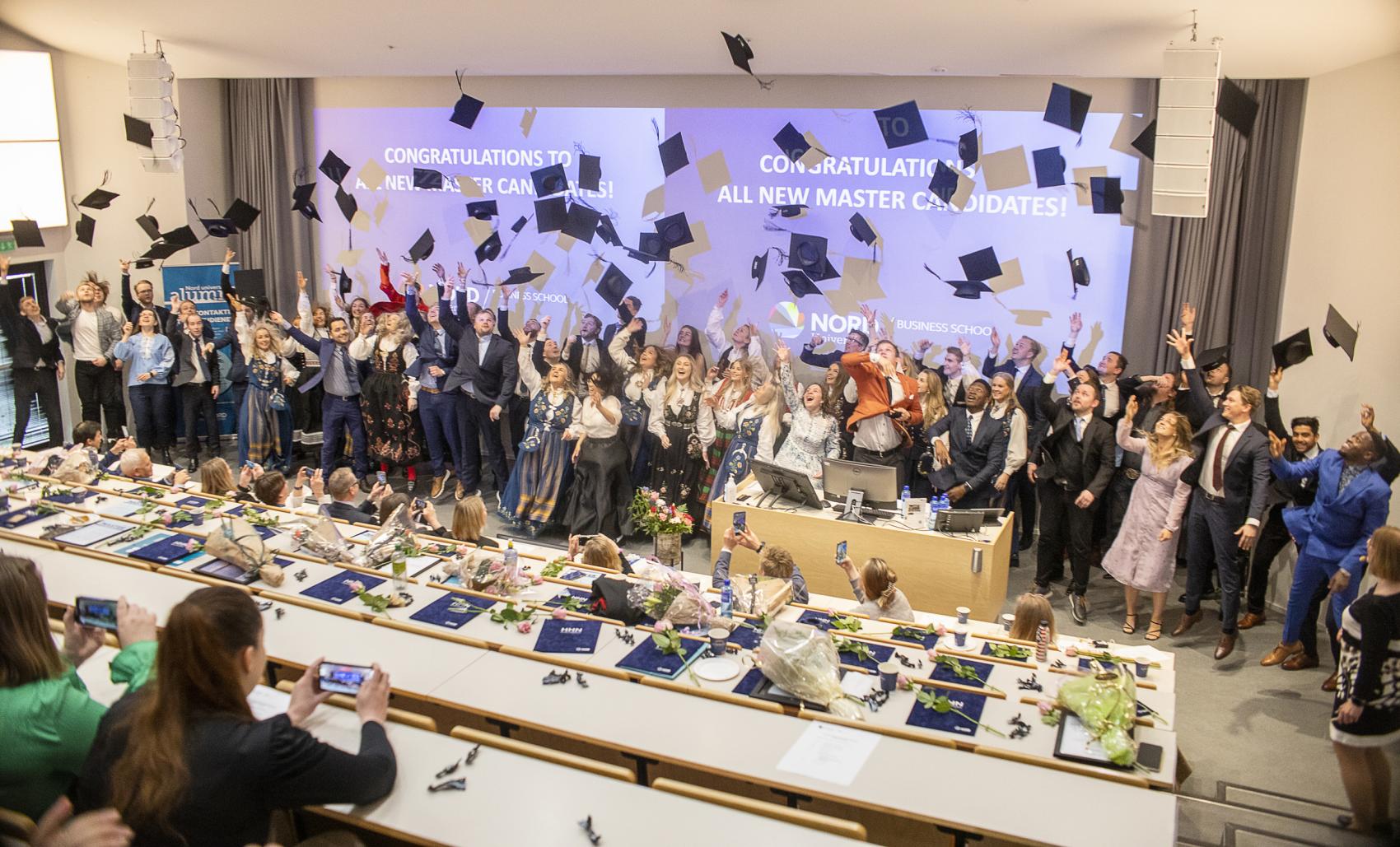 A big group of graduation students throwing their graduation hats into the air at the front of an auditorium. 