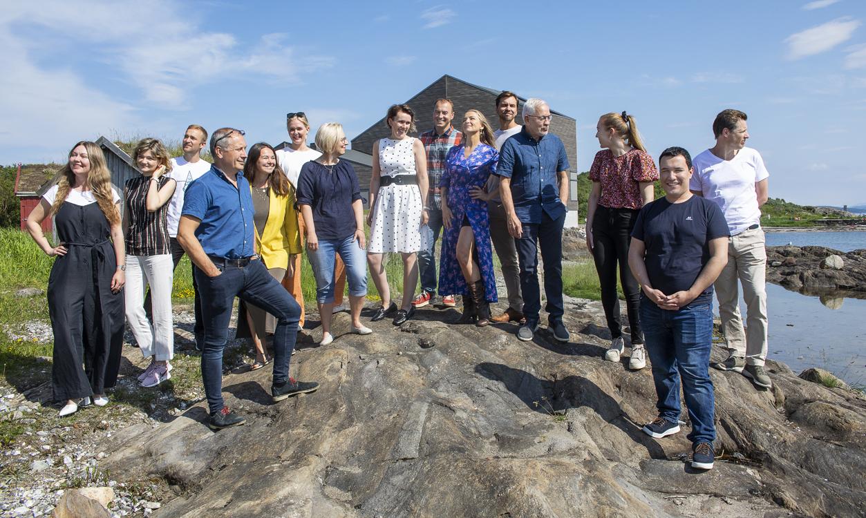 15 smiling people in summerly clothing standing next to each other on a big rock. 