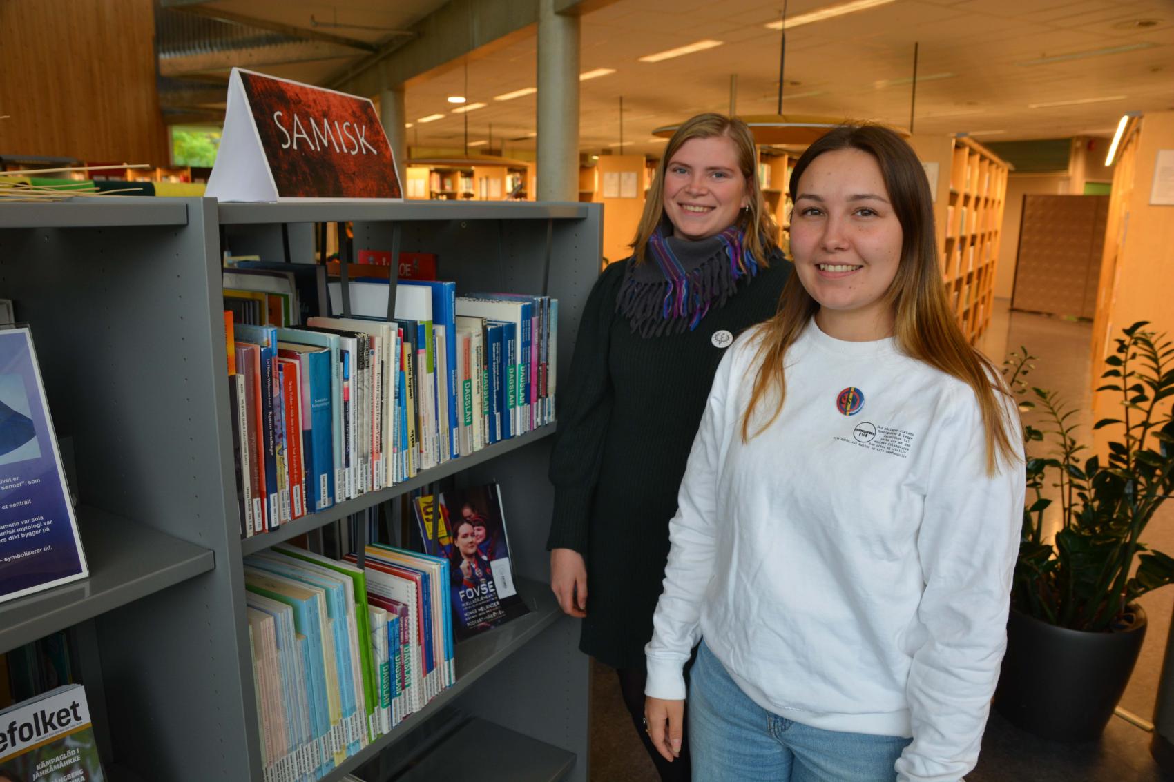 To unge samiske kvinner ved ei bokhylle i et bibliotek.