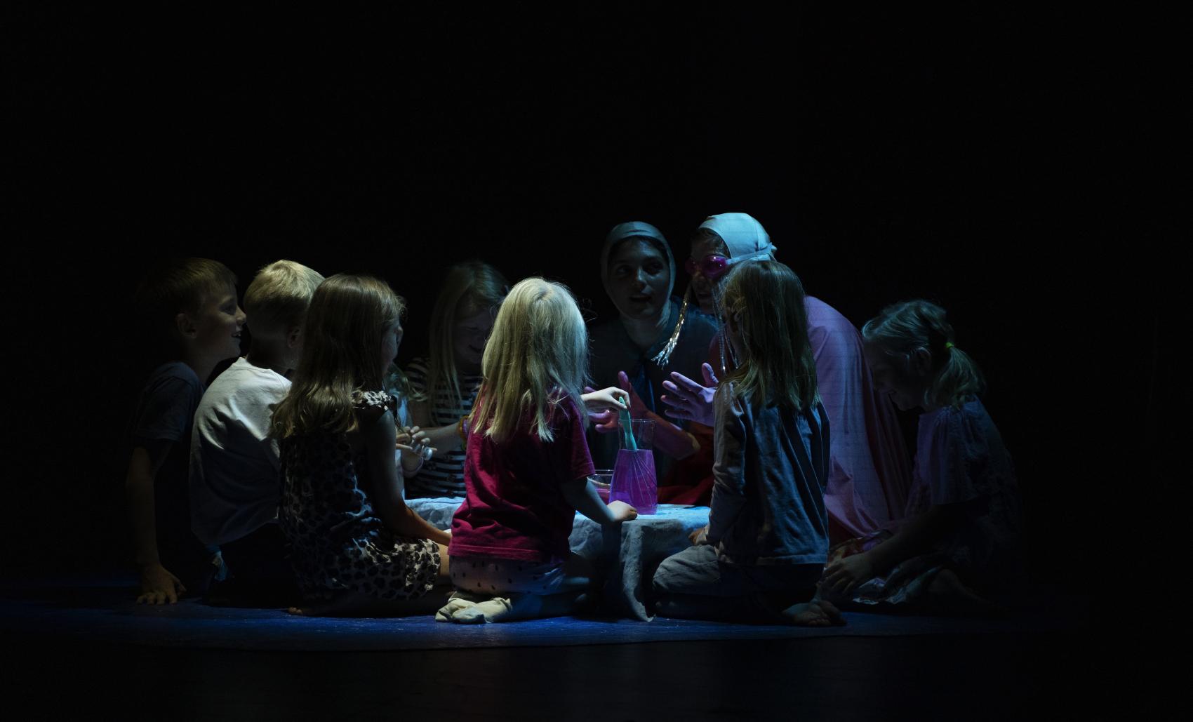 Scene fra en teaterforestilling, med mange mennesker som sitter i ring på et gulv rundt et lavt bord. Foto